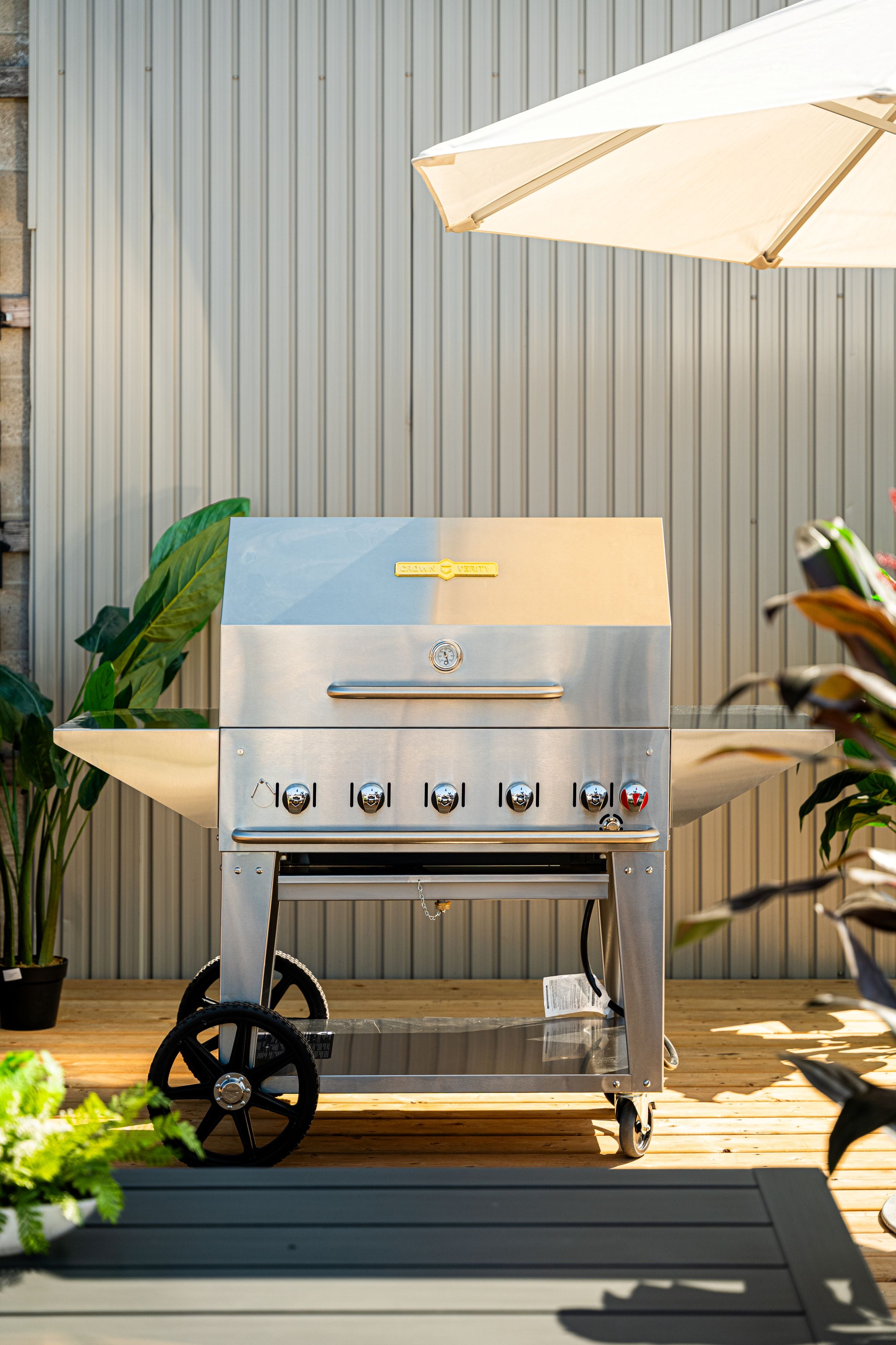 Stainless steel barbecue grill on a wooden deck with plants and an umbrella in the background.