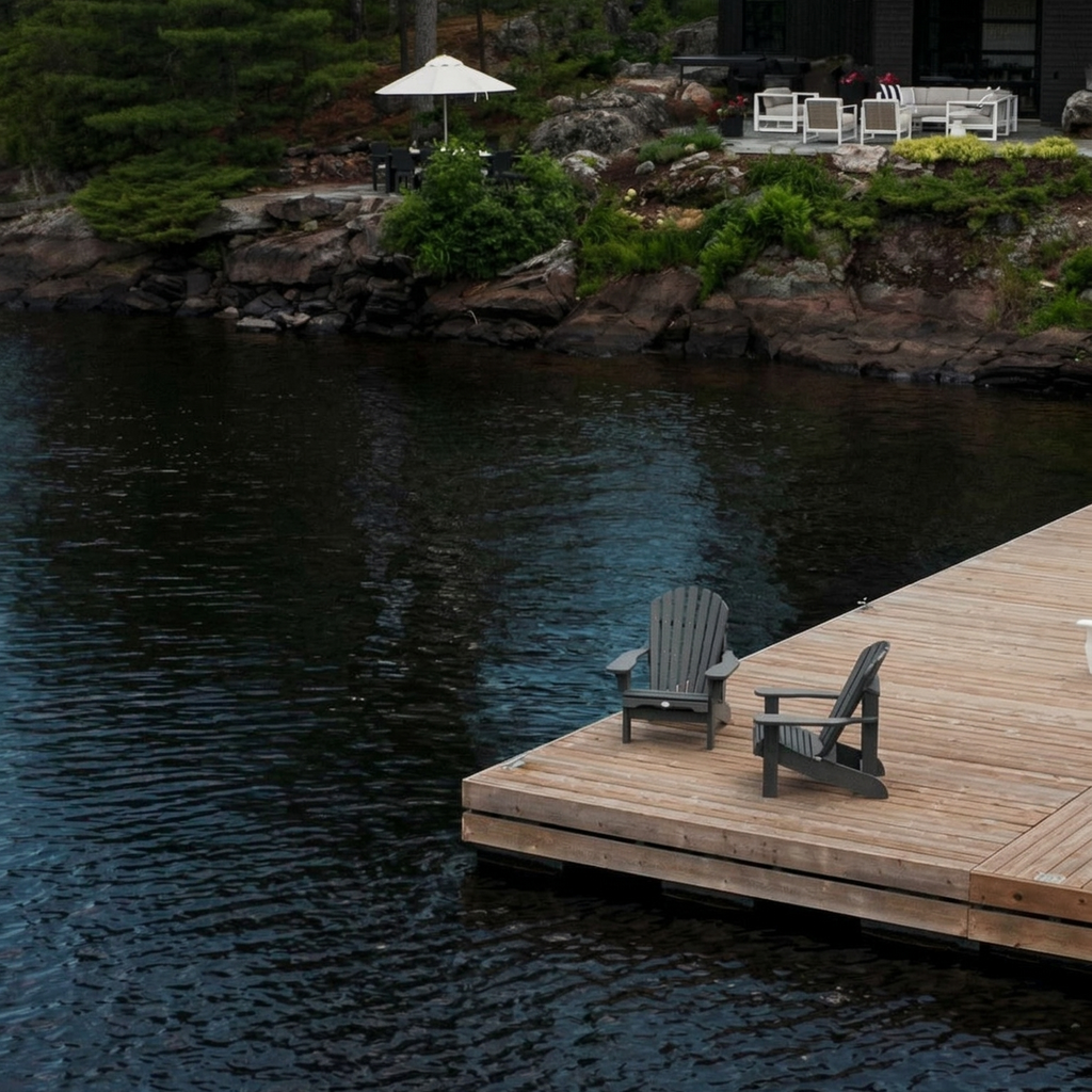 Wooden dock with chairs overlooking a calm lake surrounded by trees and a house.